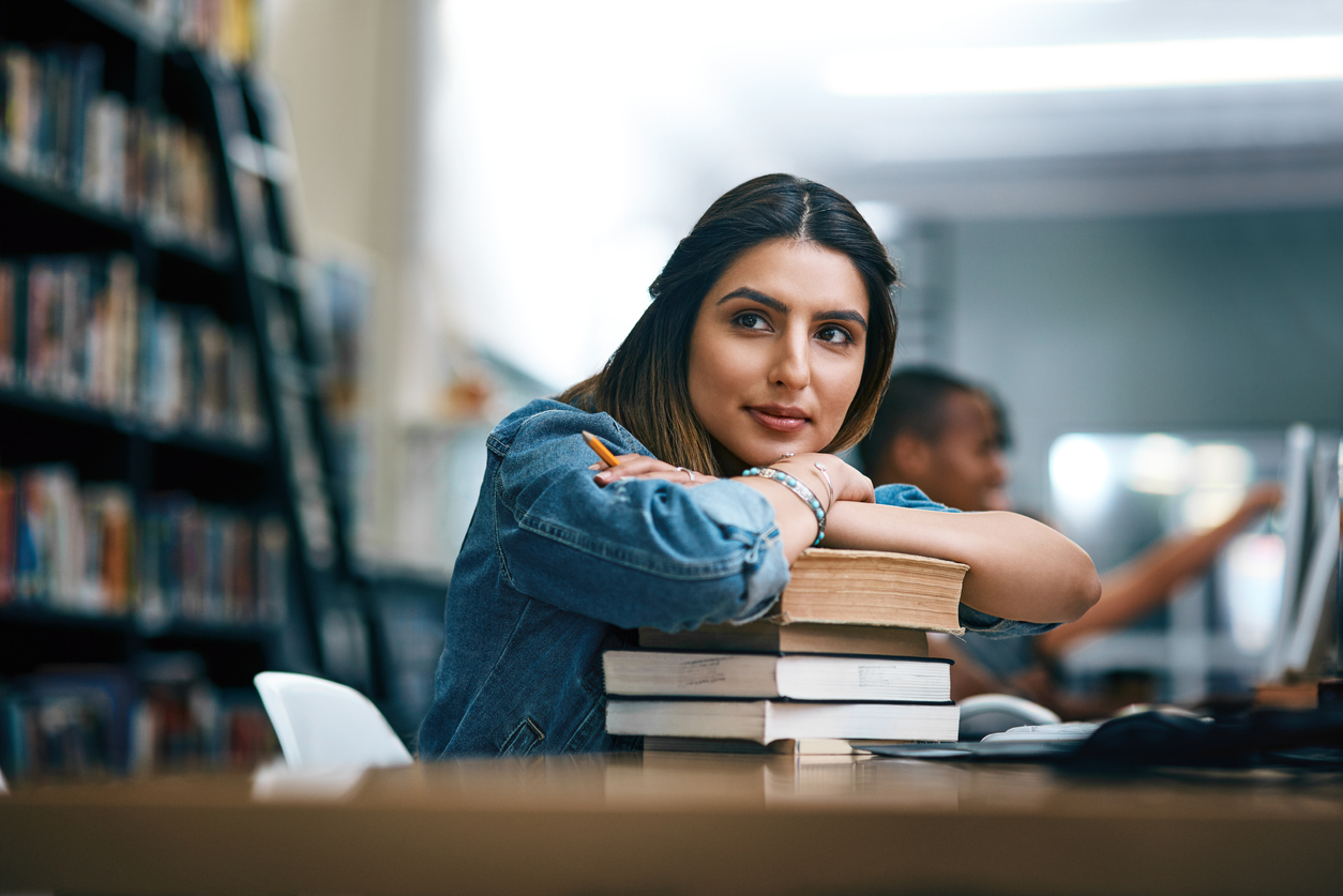 Woman in library smiling as she leans on pile of books.