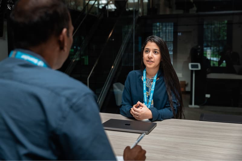 Two employees engaged in a meeting while seated across each other at a desk in an office environment.