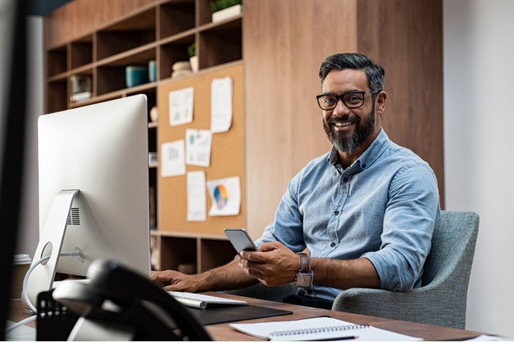man sits at desk and smiles with computer and blue shirt
