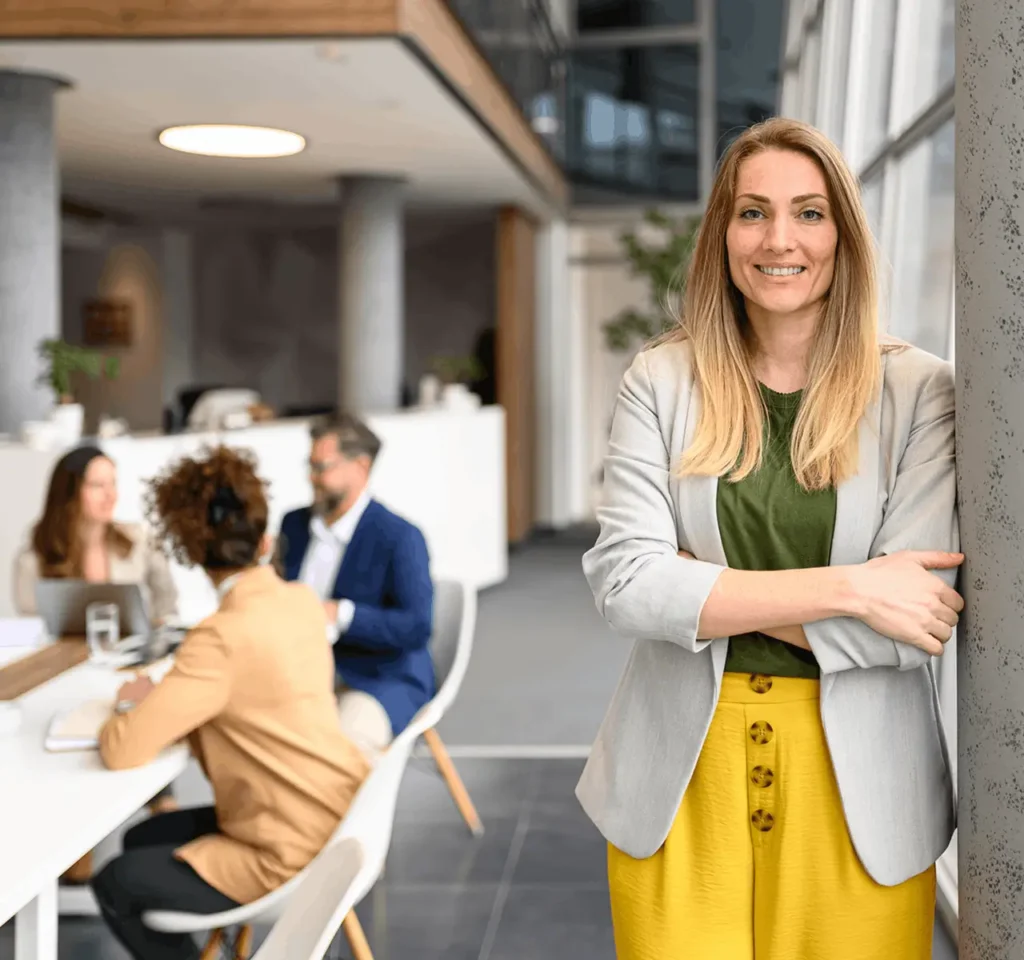 Woman smiling for the camera at her office, colleagues sitting around a desk